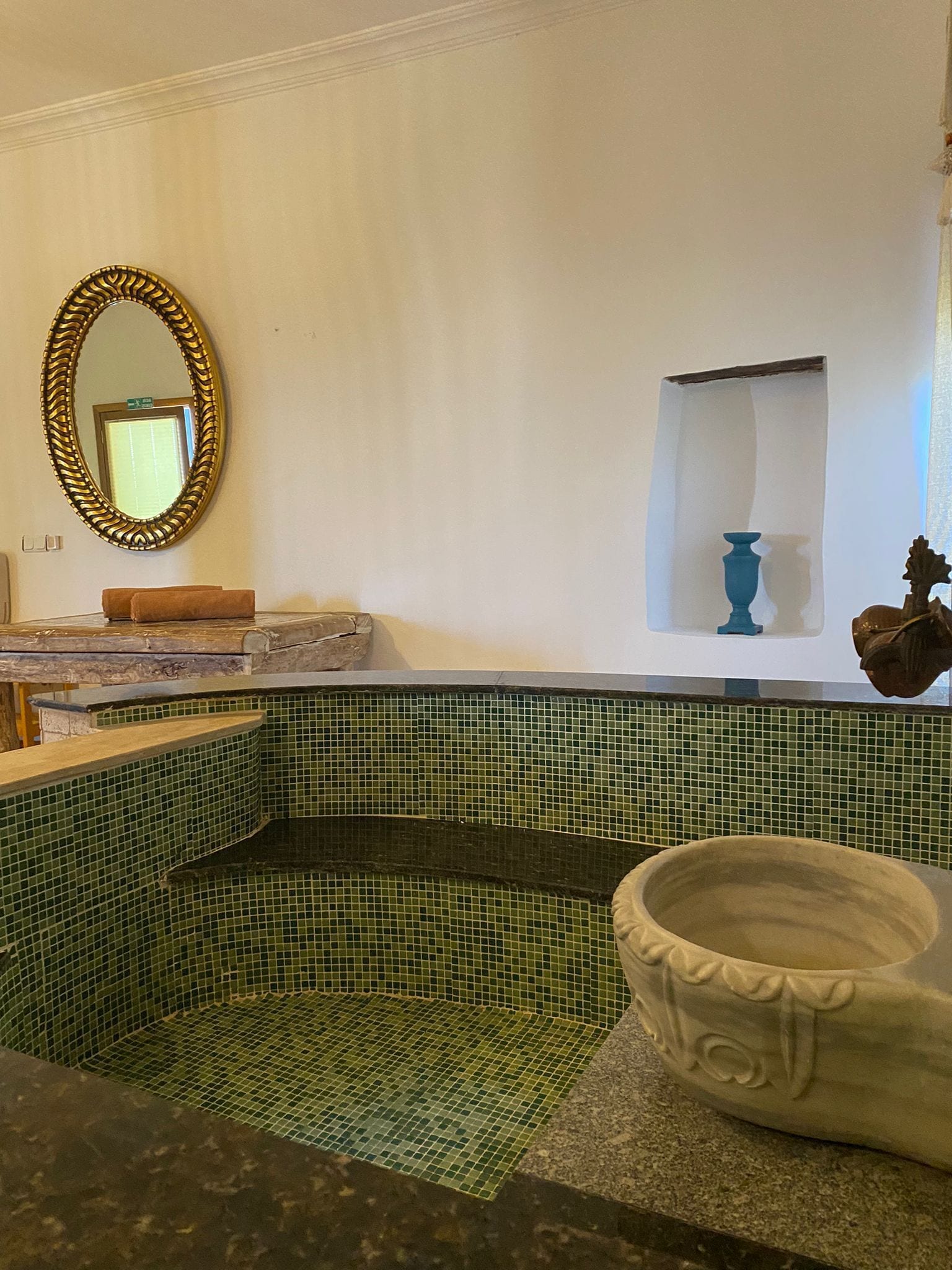 A bathroom with a mosaic tile floor, a marble countertop, a gold-framed mirror, and a small shelf with a blue vase.