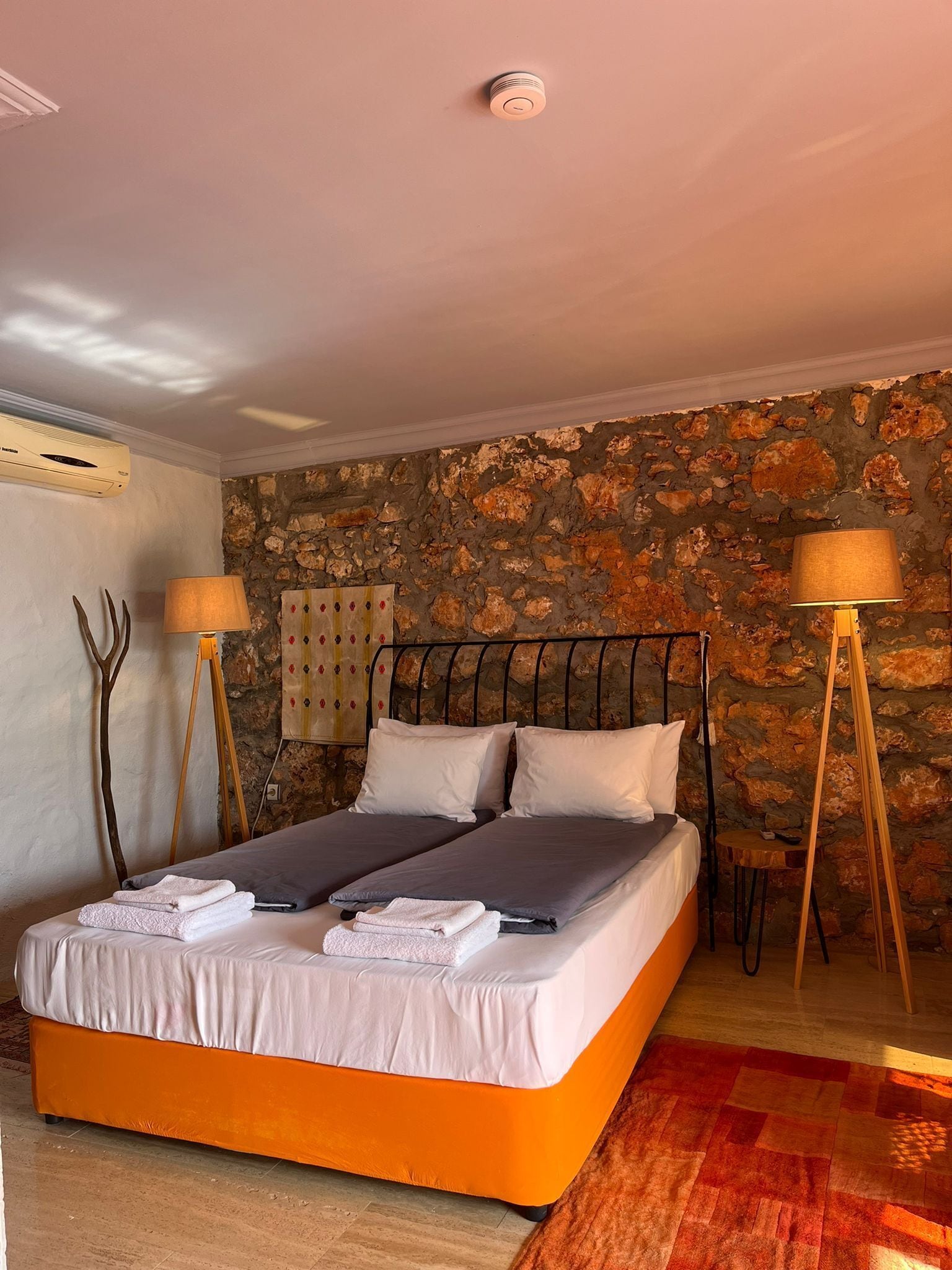 A double bed with a stone wall backdrop, a standing lamp, and a ceiling-mounted air conditioner.
