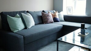 Ein modernes Wohnzimmer mit einem grauen Sofa, mehreren gemusterten Kissen und einem Glastisch.