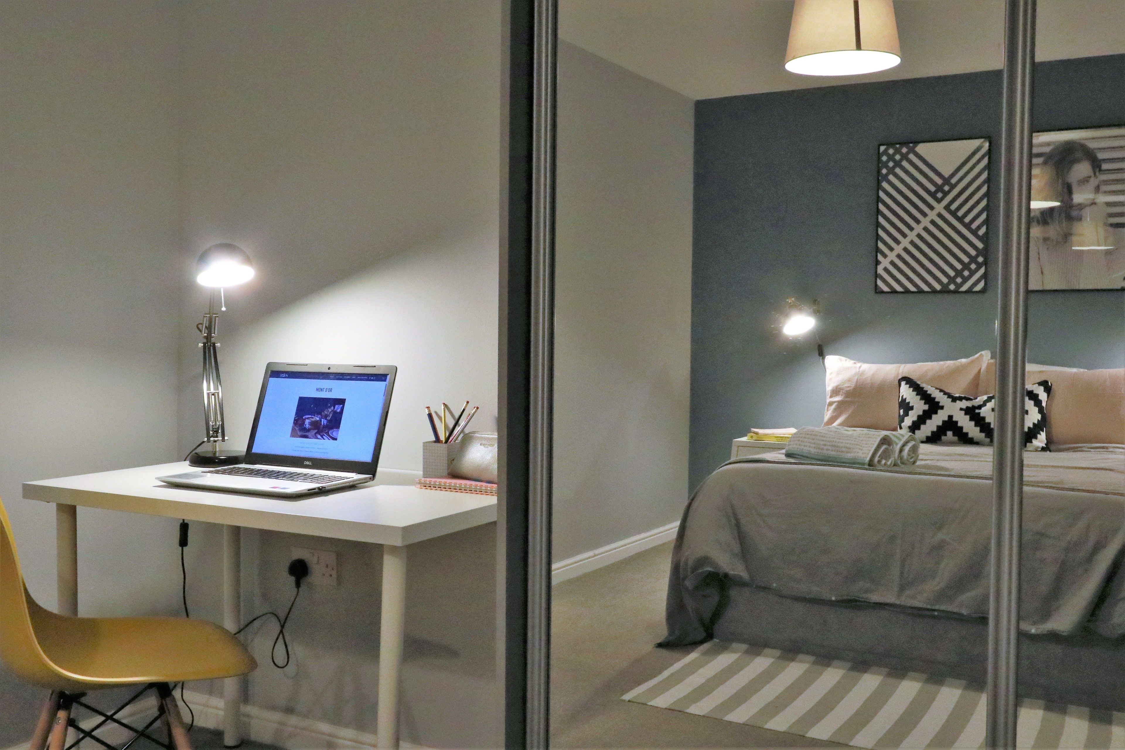 A bedroom with a bed, a desk with a laptop, a chair, and a lamp.