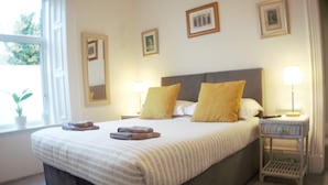 A neatly made bed with a striped bedspread and two pillows, a bedside table with a lamp, and a window with a plant. - Barns Serviced Accommodation (Ayr)