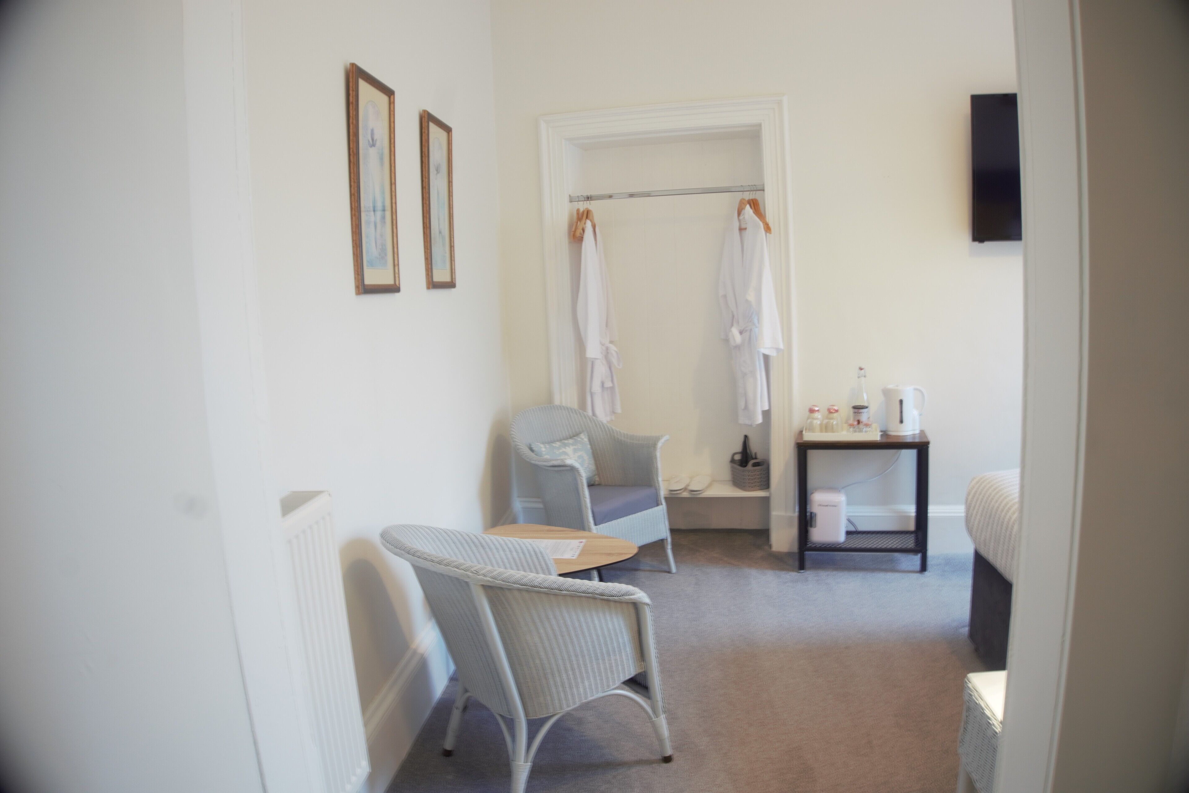 Habitación de hotel con zona de estar, una mesita, un radiador, un perchero y un televisor montado en la pared.
