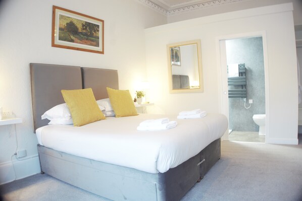 A neatly made bed with pillows, a bedside table with a lamp, a framed picture on the wall, and a bathroom visible through an open door. - Barns Serviced Accommodation (Ayr)