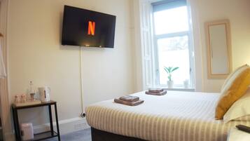 A hotel room with a bed, a TV mounted on the wall, a small table with a kettle, and a window with a plant.