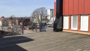 A wooden deck with a metal fence, a red building with white shutters, and a clear sky. - Hotell Gustaf (Götene)