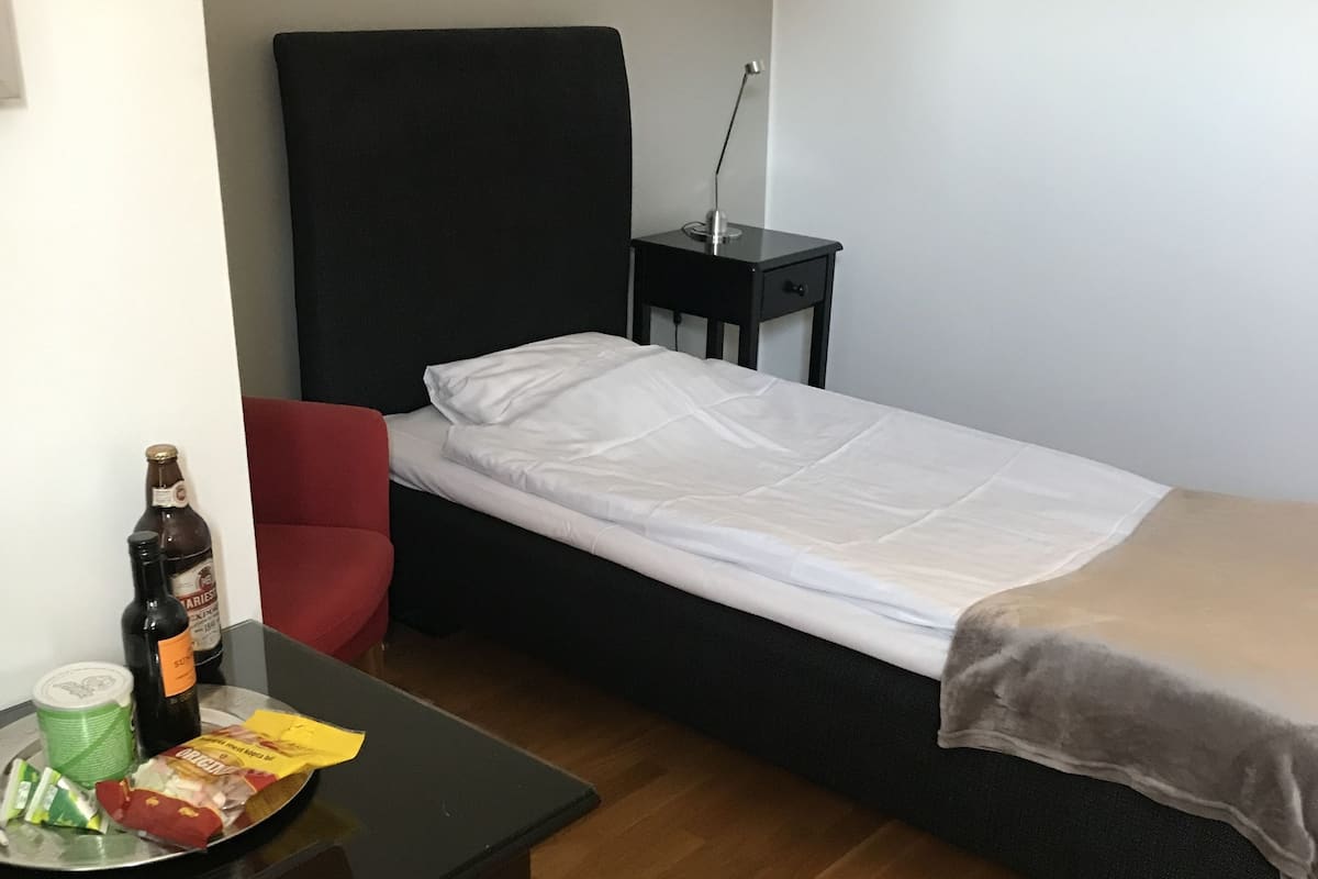 A small, neatly arranged room with a single bed, a small table with snacks and drinks, and a chair.