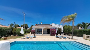 Piscina con sillones de descanso y un patio frente a una casa con techo de tejas y ventanas arqueadas.