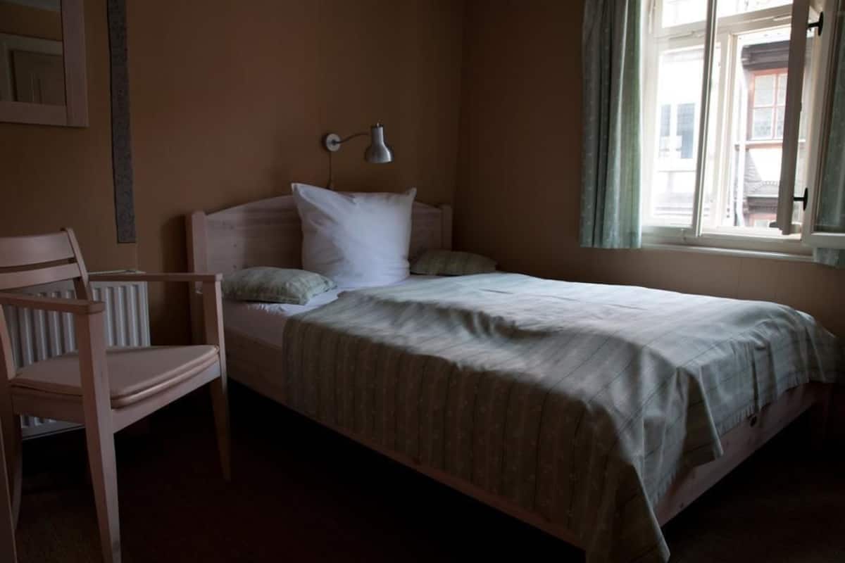 Ein Einzelbett mit Kopfteil, eine Nachttischlampe und ein Sessel in einem Zimmer mit Fenster.