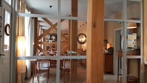 Een kamer met een grote glazen deur die leidt naar een eetgedeelte met houten stoelen en tafels. Er zijn houten balken in het plafond, en een hanglamp is zichtbaar.