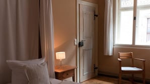 Een slaapkamer met een bed, een nachtkastje met een lamp, een houten stoel, een deur en een raam met gordijnen.
