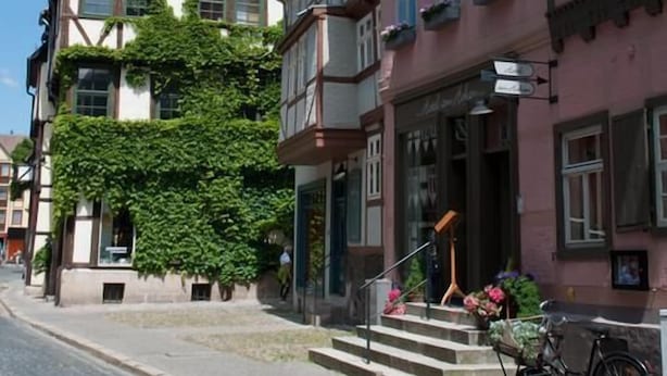 Een straat met kinderkopjes, omzoomd door traditionele Europese gebouwen, waaronder een kerk met een torenspits en een gebouw met een balkon.