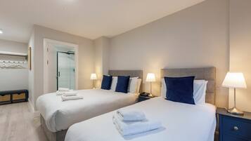 A hotel room with two beds, each with a nightstand and lamp, a small seating area with a bench, and a door leading to a bathroom.