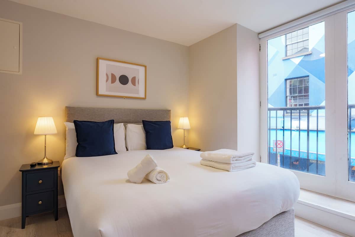 A hotel room with a large bed, two bedside lamps, a framed artwork above the bed, and a view of a building through a window.