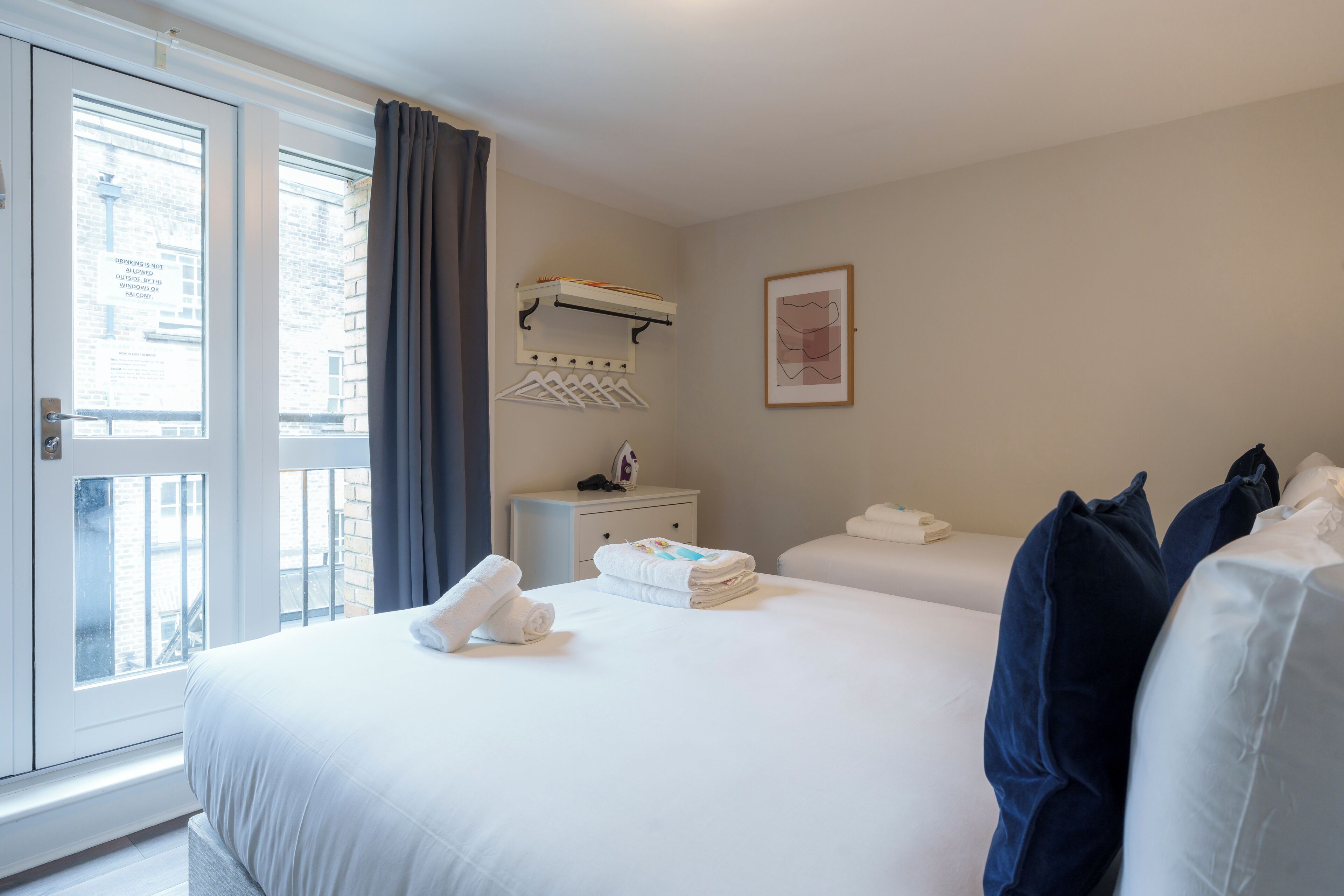 A hotel room with a bed, a bedside table, a window with curtains, a wall-mounted shelf, and a framed picture.