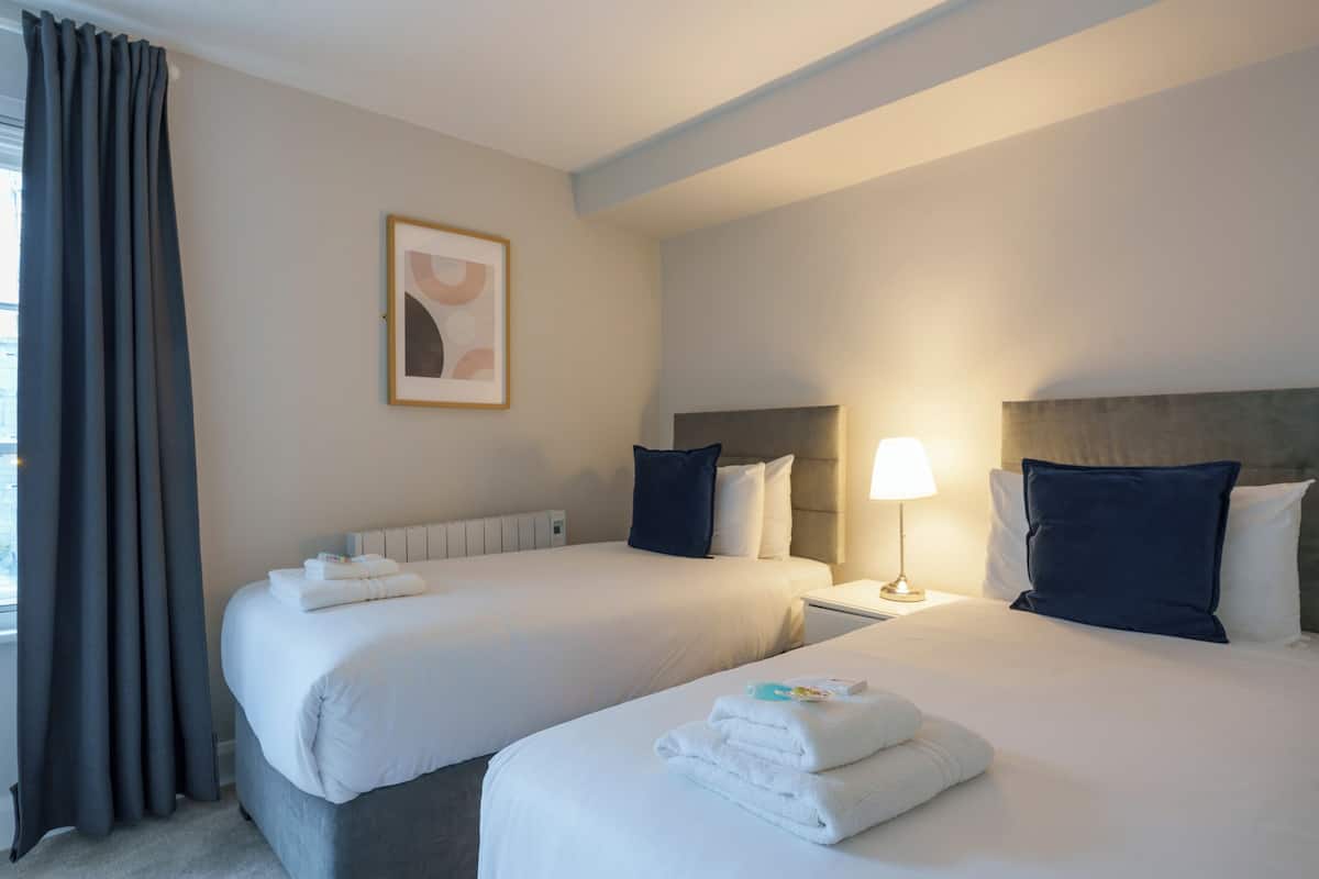 A hotel room with two beds, a nightstand with a lamp, a framed picture on the wall, and a window with curtains.