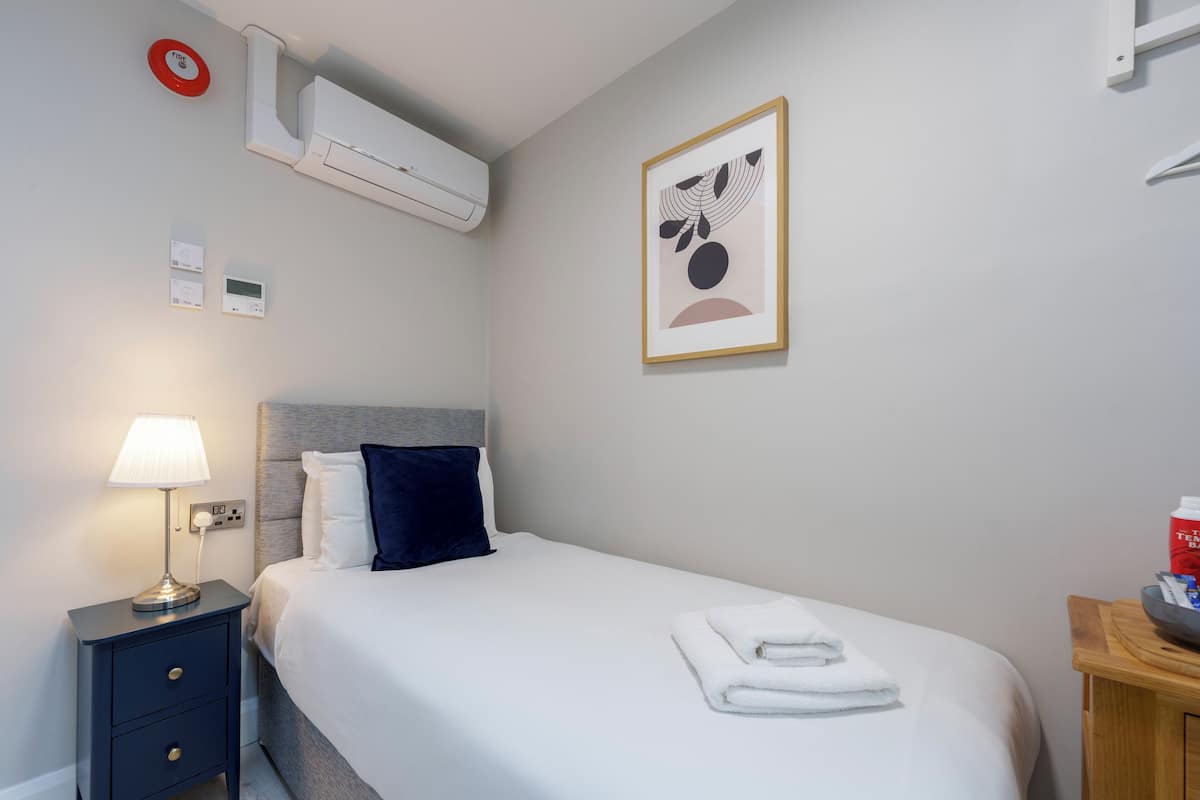 A hotel room with a bed, a bedside table with a lamp, a wall-mounted air conditioner, a framed picture, and a red fire alarm.
