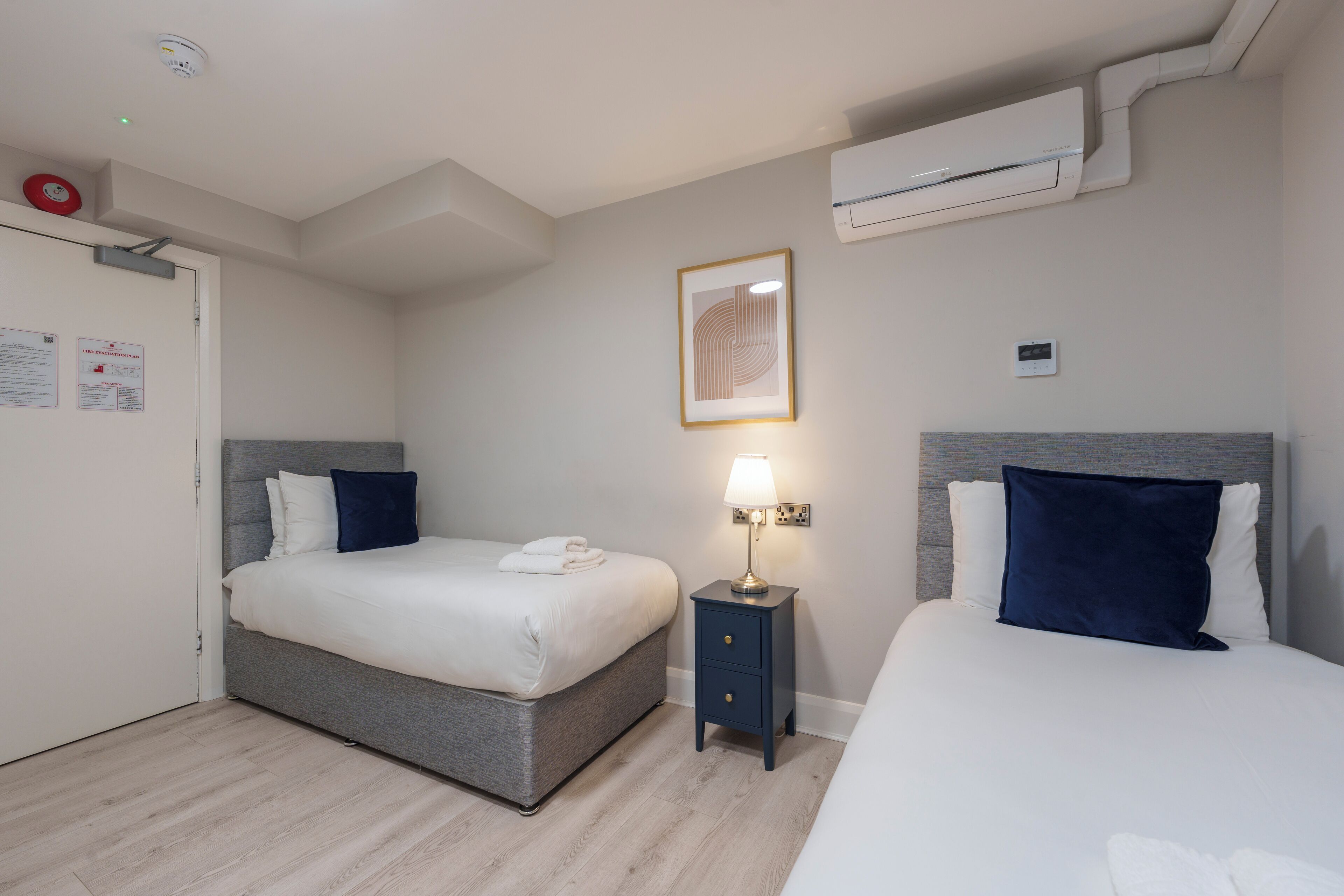 A hotel room with two beds, a bedside table with a lamp, an air conditioning unit, and a framed picture on the wall.