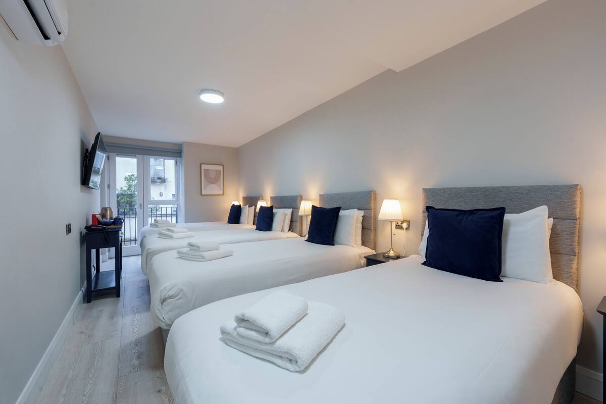 A hotel room with three beds, each with white linens and a folded towel. There is a small table with a lamp and a TV mounted on the wall.