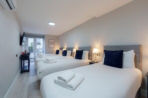 A hotel room with three beds, each with white linens and a folded towel. There is a small table with a lamp and a TV mounted on the wall.