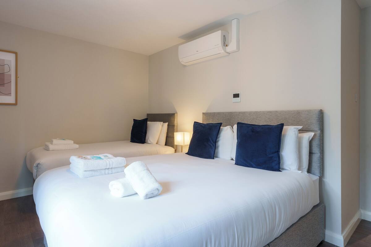 A hotel room with two beds, white linens, a wall-mounted air conditioner, and a framed picture on the wall.