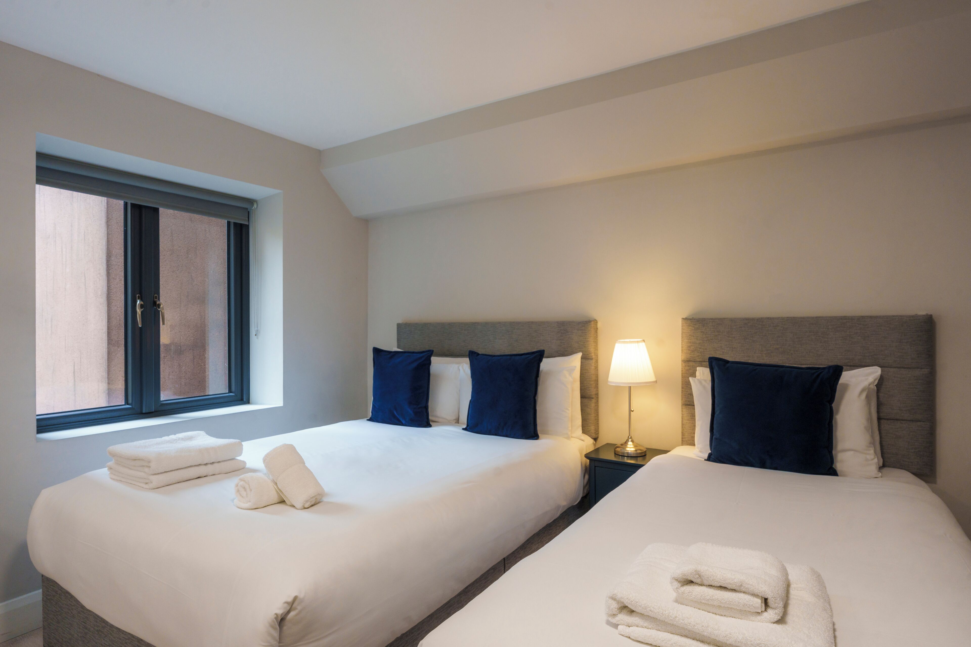 A hotel room with two beds, a window, and a nightstand with a lamp.