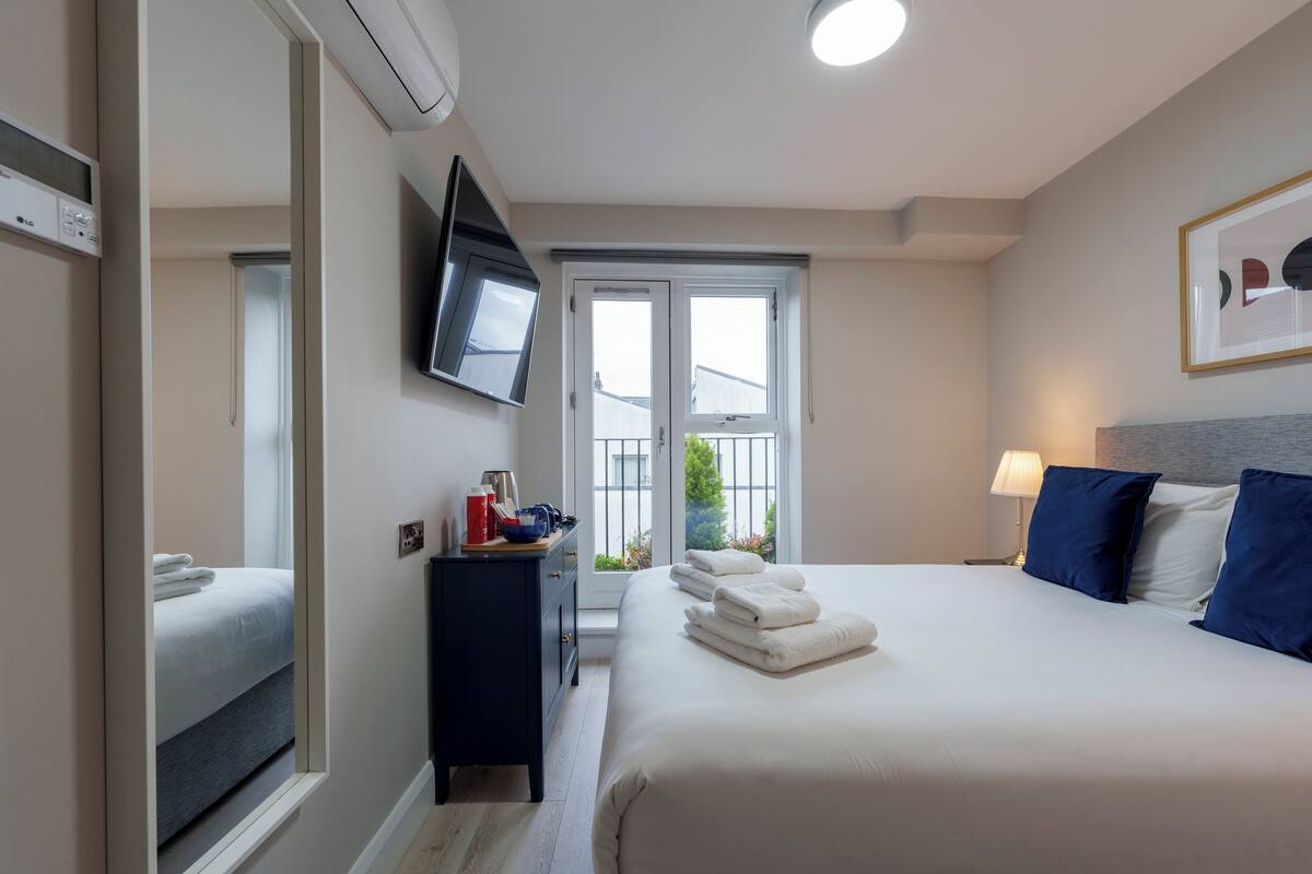 A hotel room with a bed, a TV mounted on the wall, a desk with a lamp, and a window with a view of greenery.