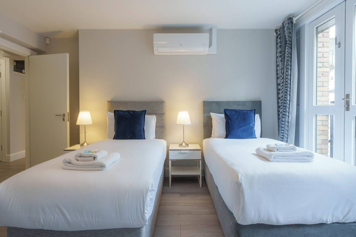 A hotel room with two beds, a nightstand with lamps, an air conditioning unit, and a view of the outside.