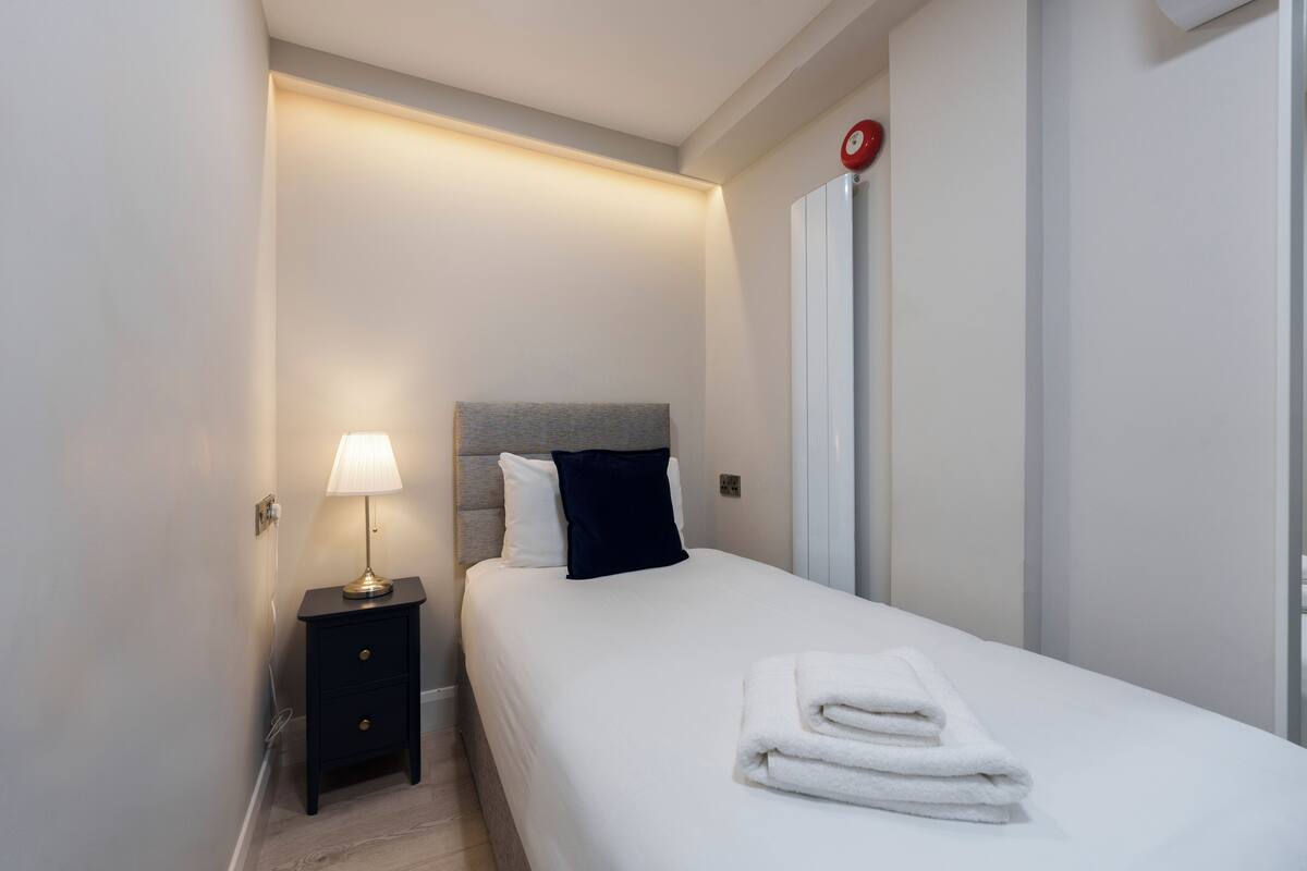 A neatly made bed with white linens and a folded towel, a bedside lamp, and a small nightstand with drawers.