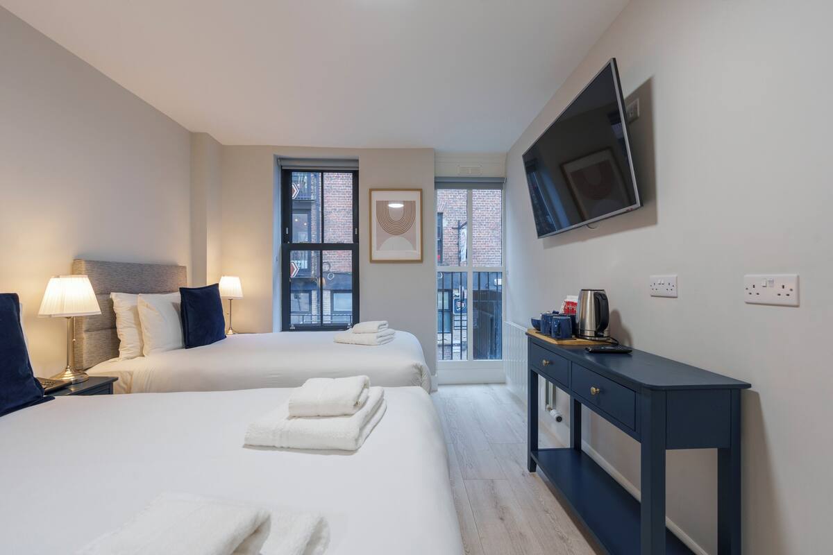 A hotel room with two beds, a TV mounted on the wall, a desk, and a window with a view of the outside.