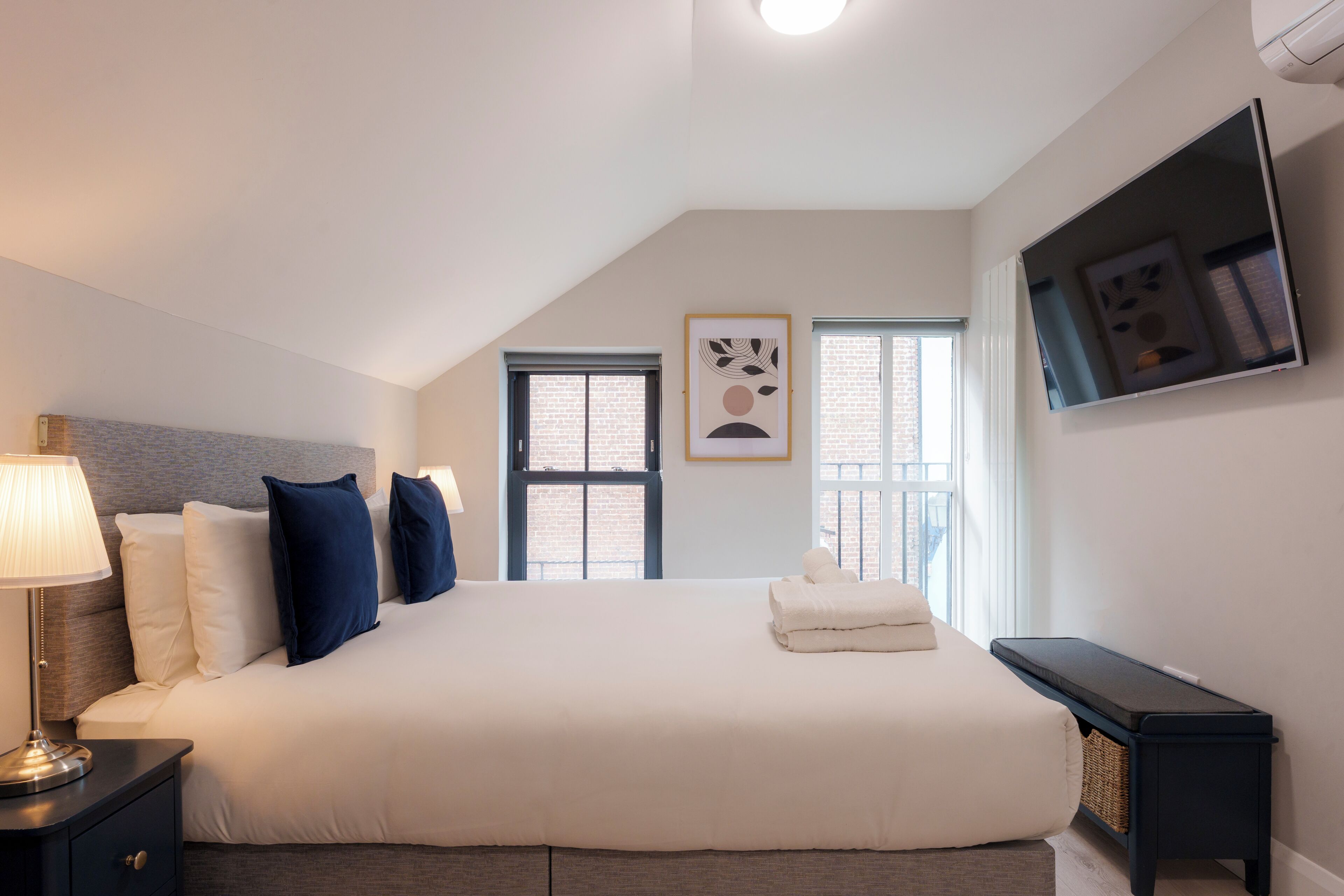 Ein Schlafzimmer mit Bett, Nachttischlampe, zwei Fenstern, einem Fernseher und einer Bank.