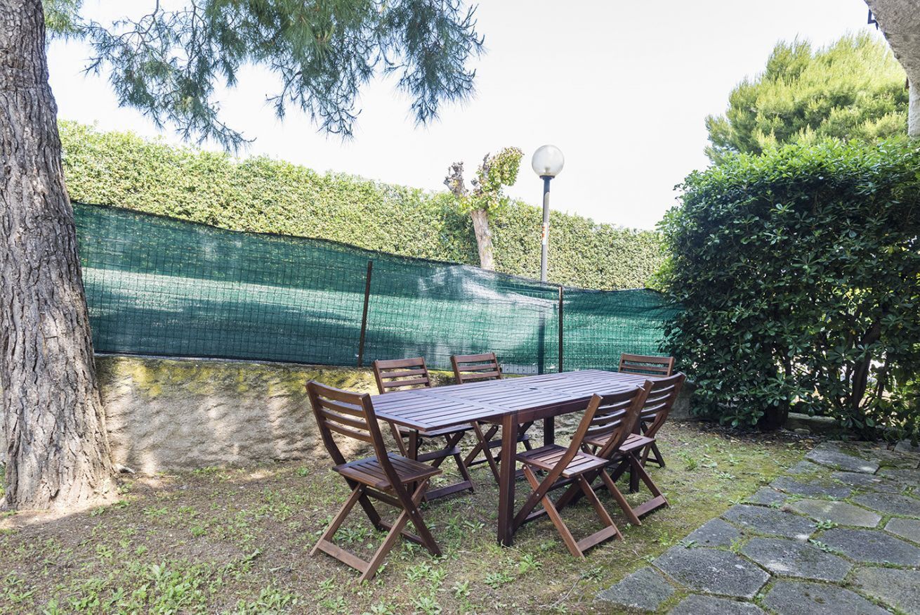 An outdoor patio with a wooden table and chairs, surrounded by a green fence and trees.