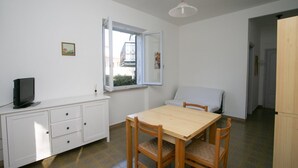 Une pièce avec une table et des chaises en bois, un meuble blanc, un téléviseur, une fenêtre donnant sur un balcon et un luminaire suspendu.