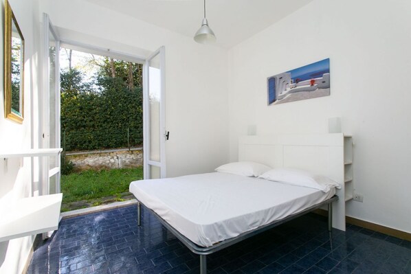 Ein Schlafzimmer mit Bett, weißen Wänden, einem Gemälde und Blick auf Grün.