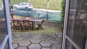 Un espace patio doté d’un sol en pierre hexagonale, de mobilier en bois et offrant une vue sur une route et une clôture, le tout visible à travers une porte vitrée.