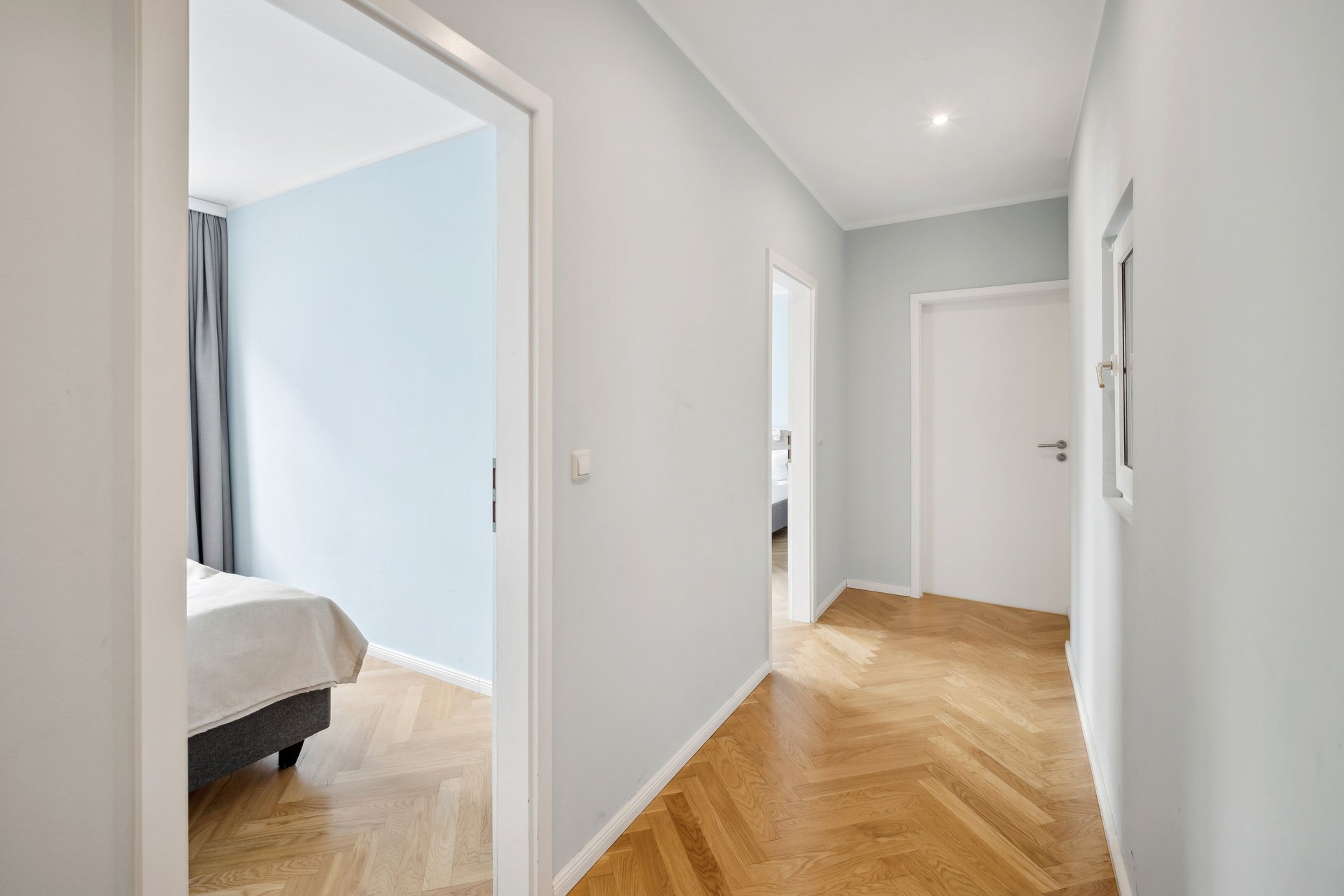 A hallway with wooden flooring and white walls, leading to a bedroom with a bed and a door.