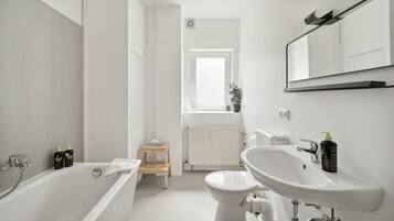 Ein modernes Badezimmer mit Badewanne, Toilette und Waschbecken. Auf der Fensterbank stehen ein kleiner Holzschemel und ein Blumentopf.