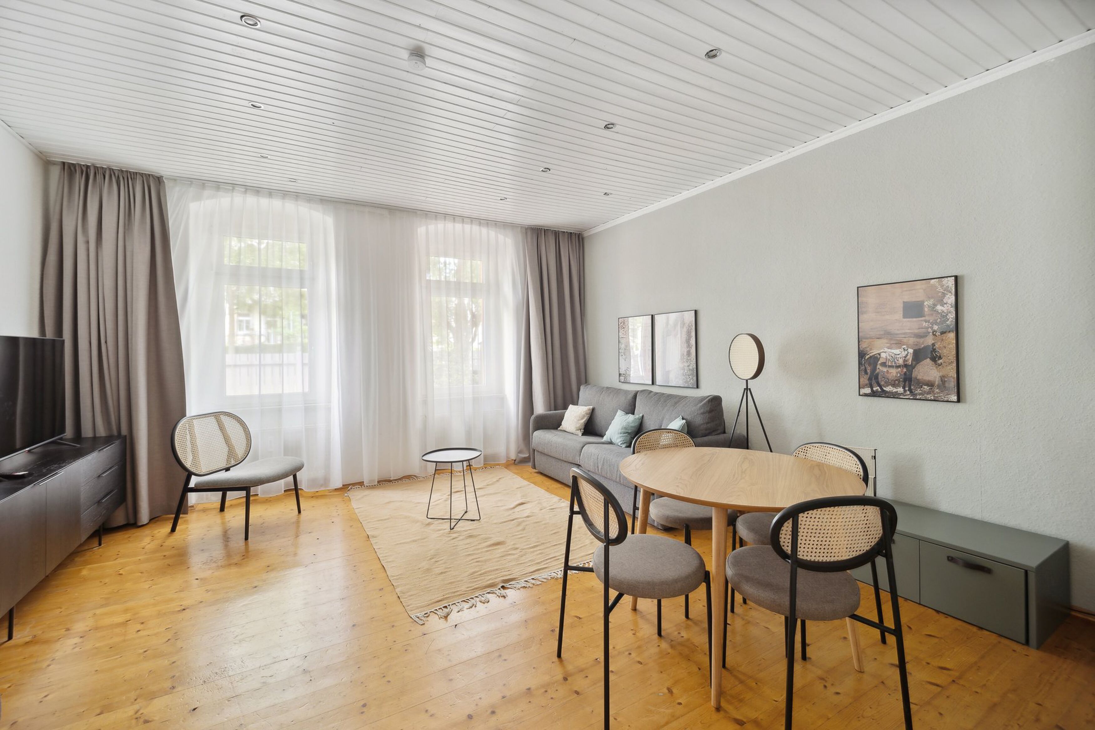 Una sala de estar moderna con un sofá, una mesa de comedor con sillas, un televisor y un amplio ventanal con cortinas.