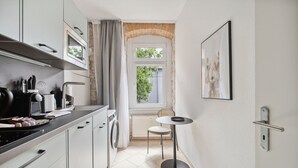 Cocina compacta con armarios blancos, microondas, hervidor y una pequeña mesa con una silla.