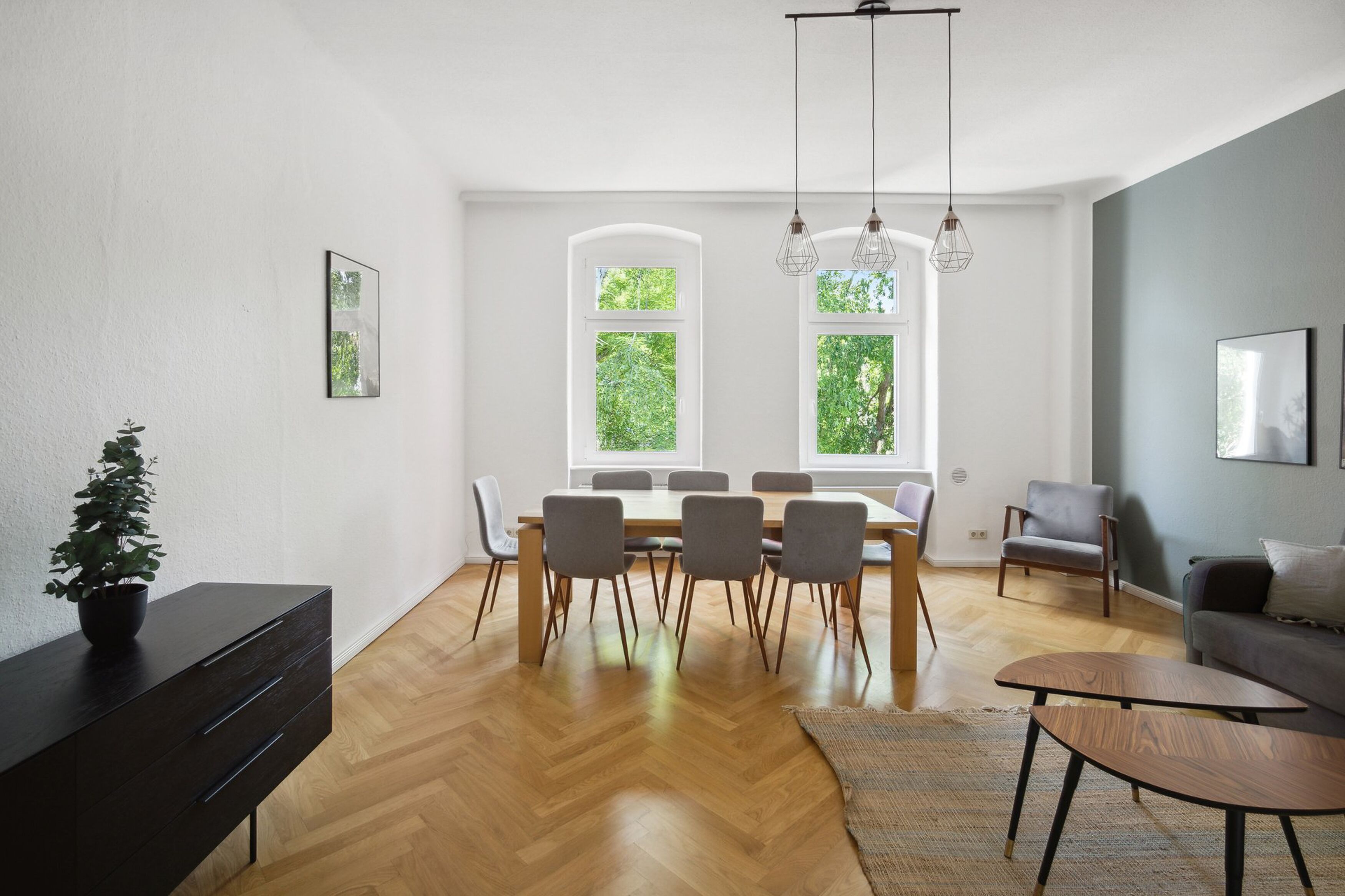 A modern living room with a dining area, large windows, and a view of greenery.