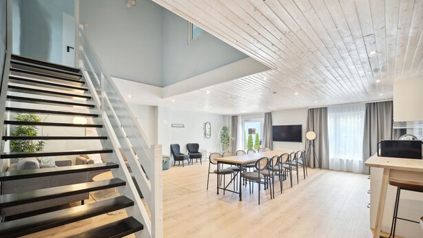 Amplia sala de estar con escalera de madera, una mesa de comedor con sillas y una zona de cocina con televisor.