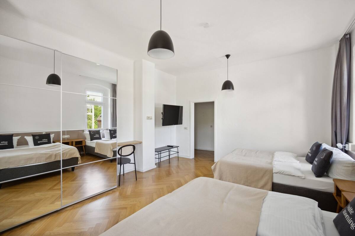 A modern hotel room with a large mirror, a flat-screen TV, a wooden floor, and a bed with a headboard.