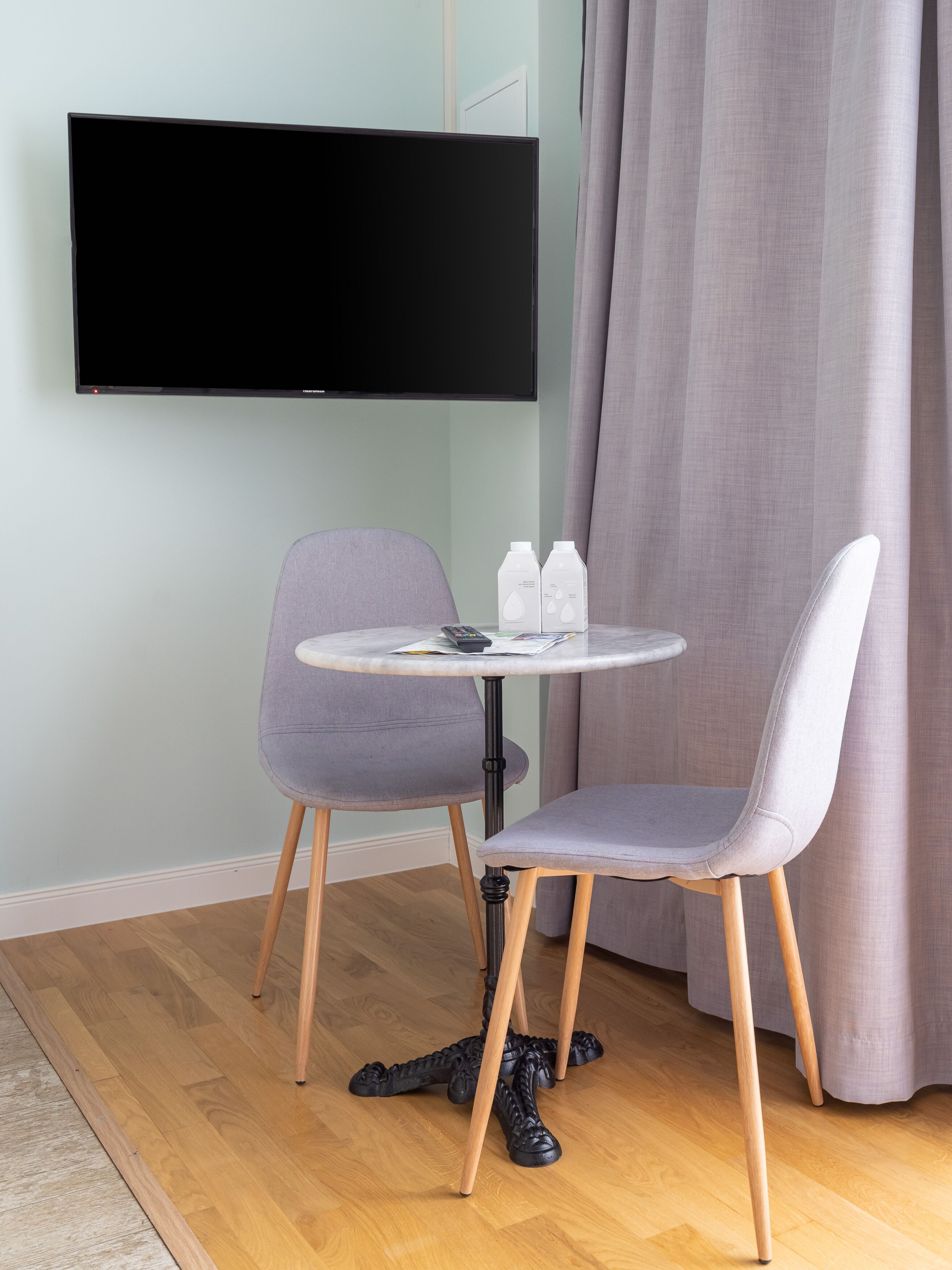 Una habitación moderna con un televisor de pantalla plana montado en la pared, una mesa redonda con dos sillas grises y un par de zapatos negros en el suelo.