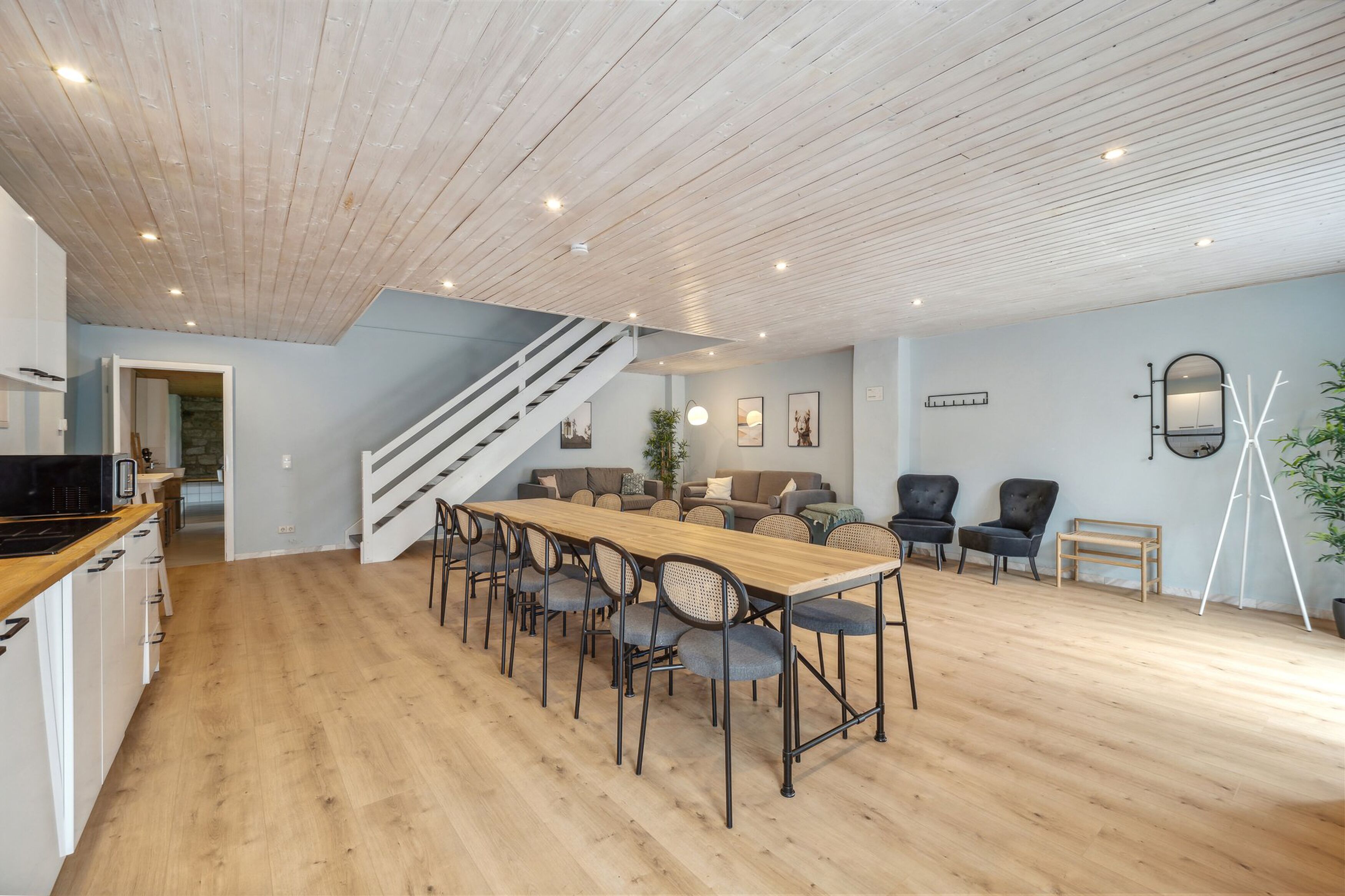 A modern interior with a wooden dining table, chairs, a staircase, and a living area with sofas and chairs.