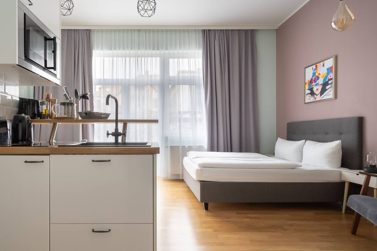 A modern hotel room with a kitchenette, a bed, a bedside table, and a window with curtains.