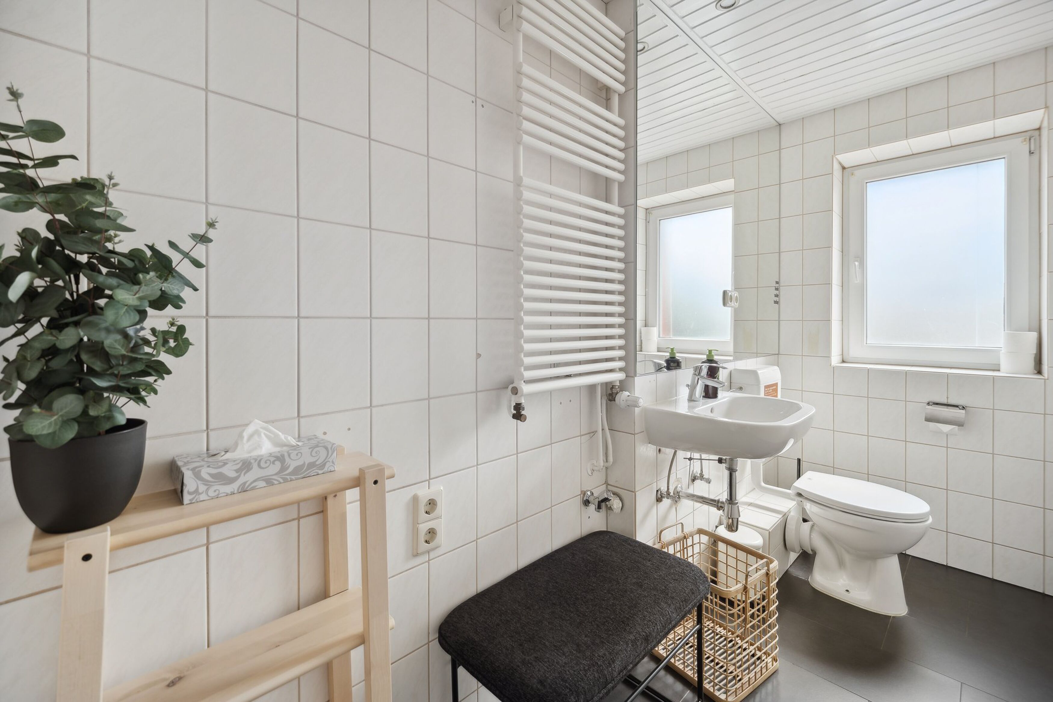 A modern bathroom with white tiles, a radiator, a sink, a toilet, a bench, and a potted plant.