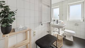 A modern bathroom with white tiles, a radiator, a sink, a toilet, a bench, and a potted plant.