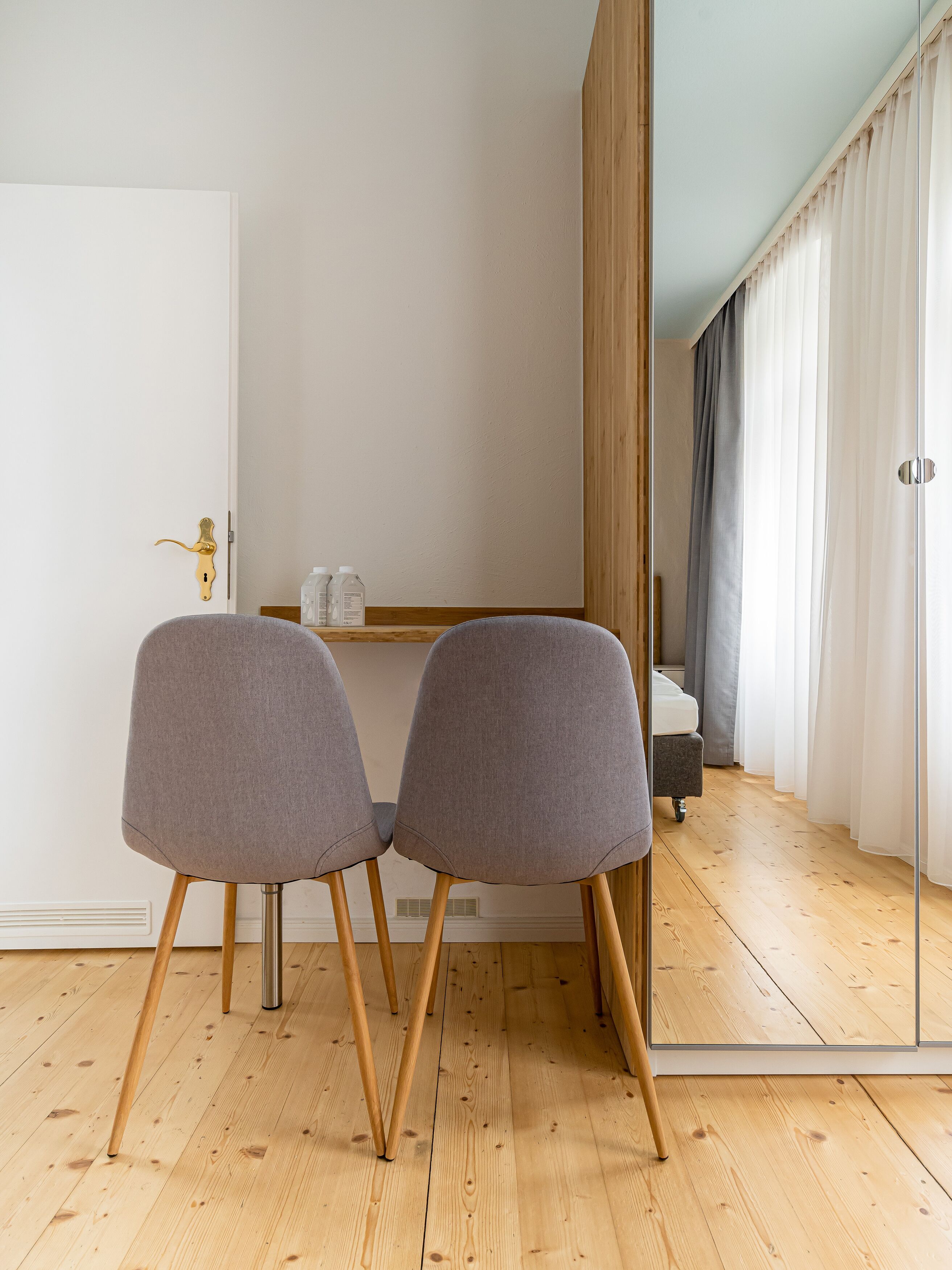 A modern room with a wooden floor, a white door, two grey chairs, a small wooden table with a bottle, and a mirror reflecting a bed and curtains.