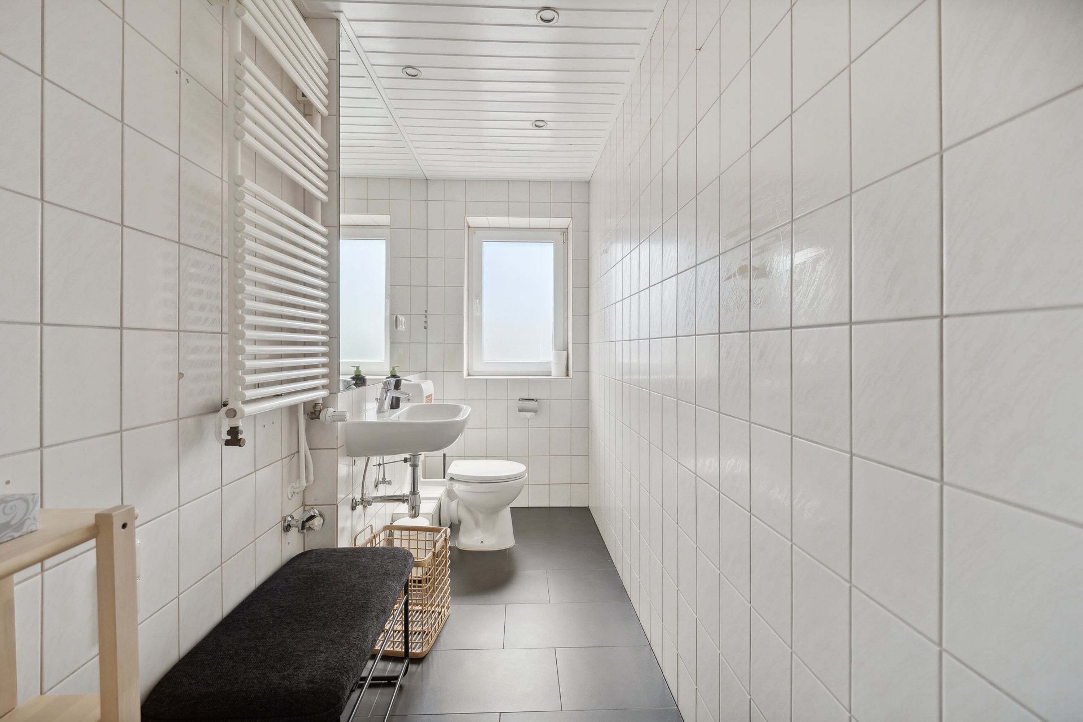 Ein schmales Badezimmer mit weißen Fliesen, einer Toilette, einem Waschbecken und einer Sitzbank.