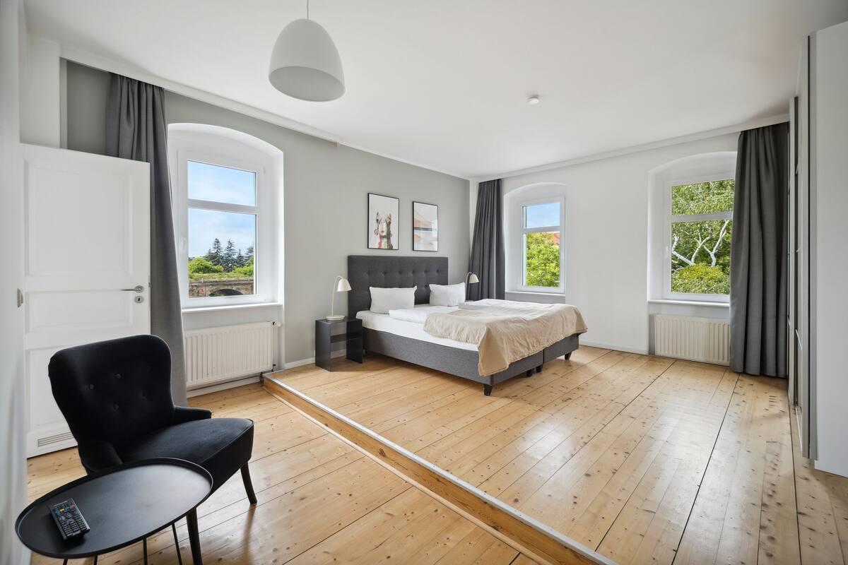 A modern bedroom with a large bed, a chair, a small table, and two windows with curtains.