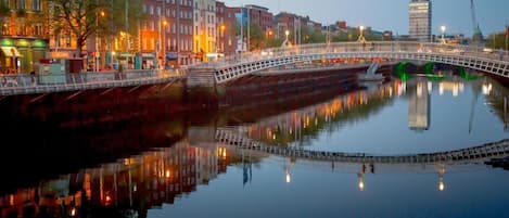 A bridge over a calm river with buildings on either side, a clear sky, and a reflection of lights on the water.